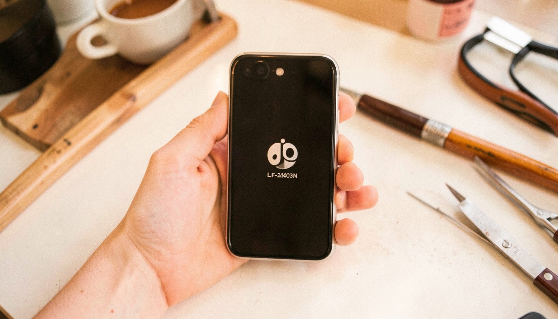 Jio Phone LF-2403N mudel käes hoides, ekraanil taaskäivituse logo. Taustal on hele töölaud koos tööriistadega.
