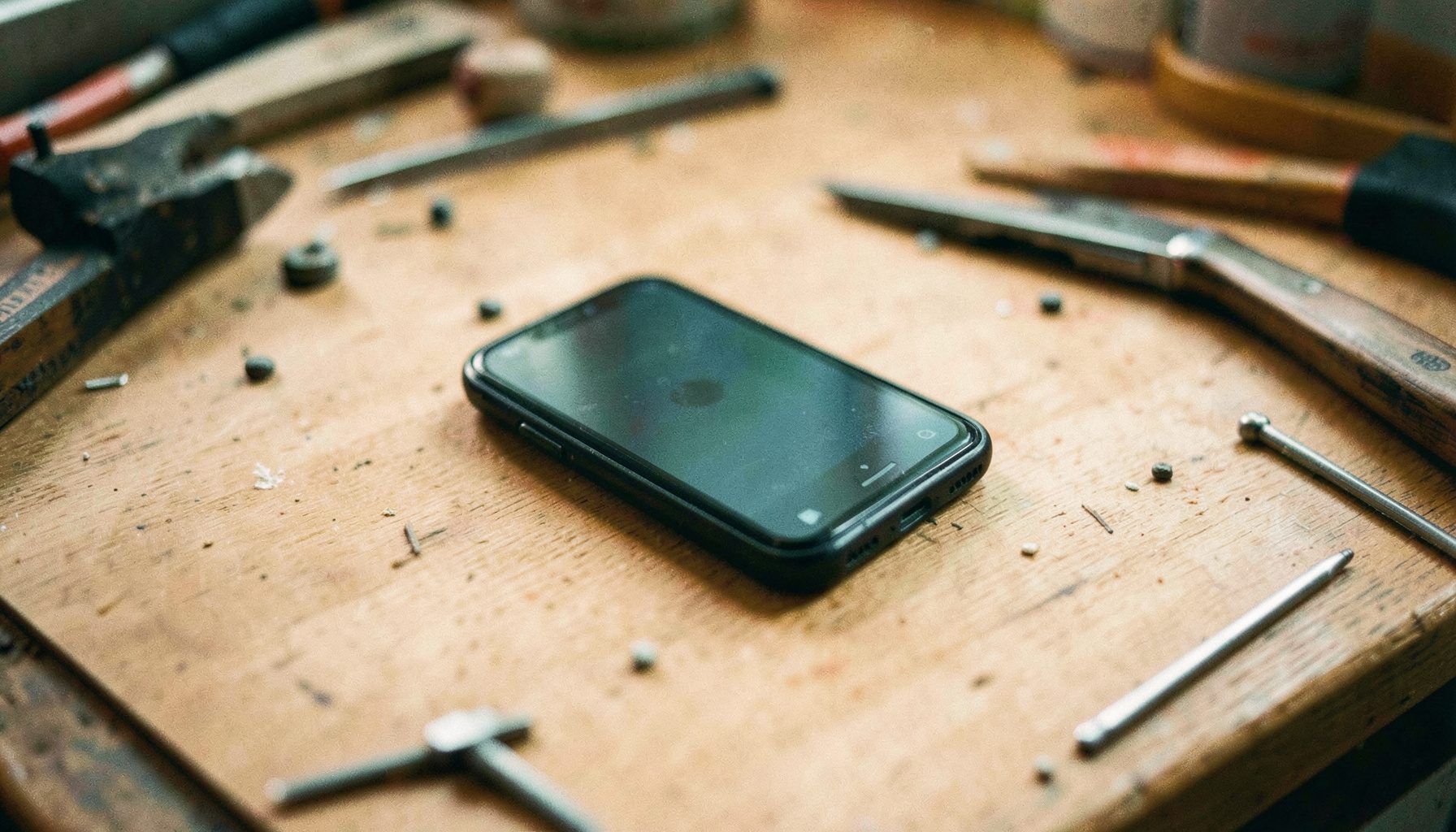 Lähivõte lahti võetud mobiiltelefoni trükkplaadist töölaual, ümber väikesed kruvid ja tööriistad. Soe, hämar valgustus rõhutab vase ja rohelise toone.