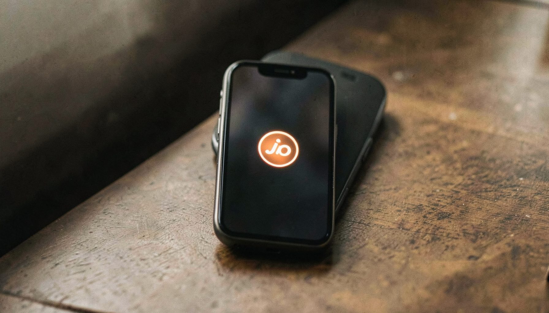 Lähivõte Jio feature phone'ist, mille ekraanil kuvatakse taaskäivituse logo. Telefon on asetatud neutraalsele hallile pinnale, pehme loomuliku valgusega. Domineerivad tumehallid ja mustad toonid koos ekraani sinaka helgiga.